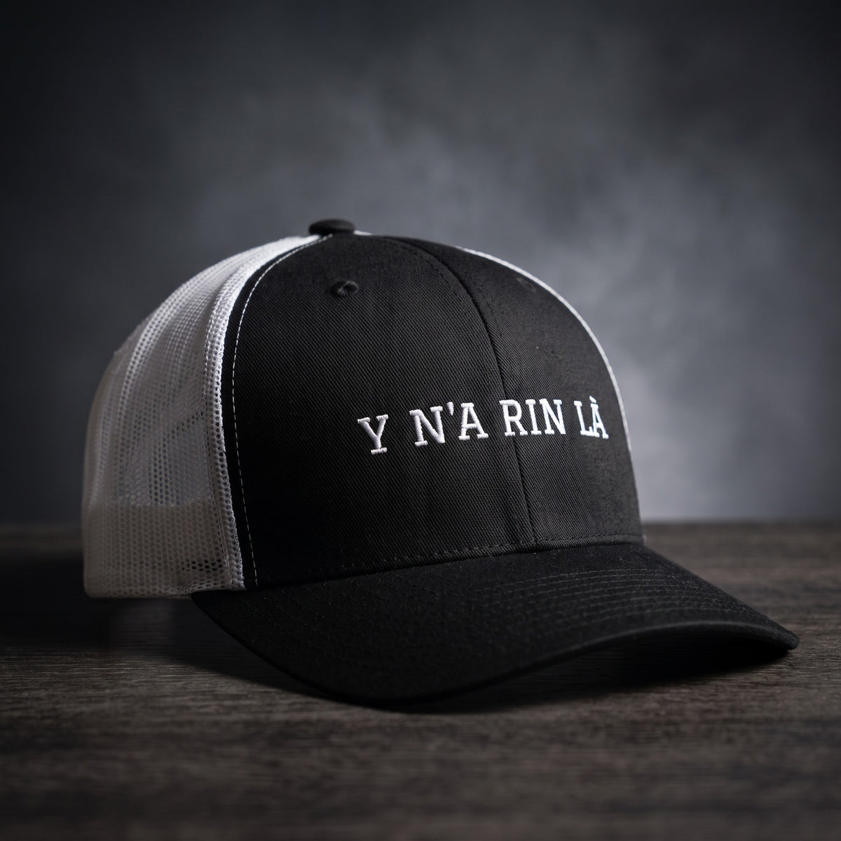 Casquette trucker noire et blanche brodée « Y N'A RIN LÀ » en blanc, posée sur un bois sombre