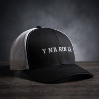 Casquette trucker noire et blanche brodée « Y N'A RIN LÀ » en blanc, posée sur un bois sombre