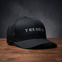 Casquette trucker noire brodée « Y N'A RIN LÀ » en blanc, posée sur un bois sombre
