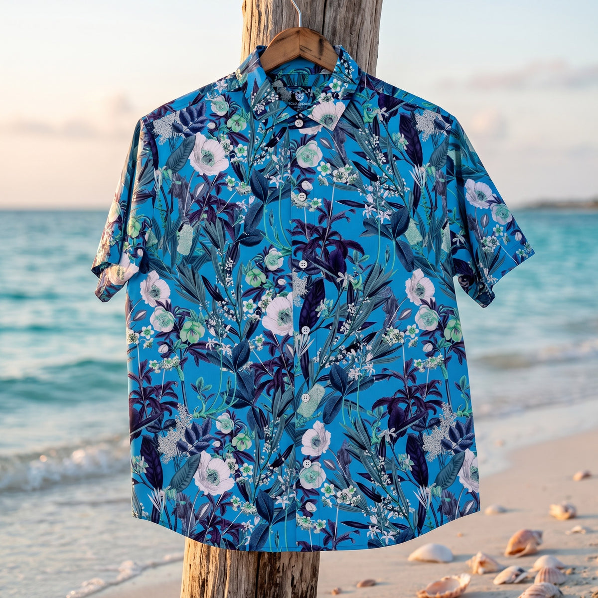 Chemise homme à manches courtes bleue à fleurs blanches et feuillage marine suspendue sur un cintre, à un piquet en bois flotté sur une plage tranquille avec coquillages, lumière douce du matin - CHT'M Boutique