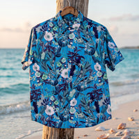 Chemise homme à manches courtes bleue à fleurs blanches et feuillage marine suspendue sur un cintre, à un piquet en bois flotté sur une plage tranquille avec coquillages, lumière douce du matin - CHT'M Boutique