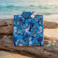 Chemise homme à manches courtes bleue à fleurs blanches et feuillage marine pliée sur une planche en bois flotté sur une plage avec coquillages, lumière douce du matin - CHT'M Boutique