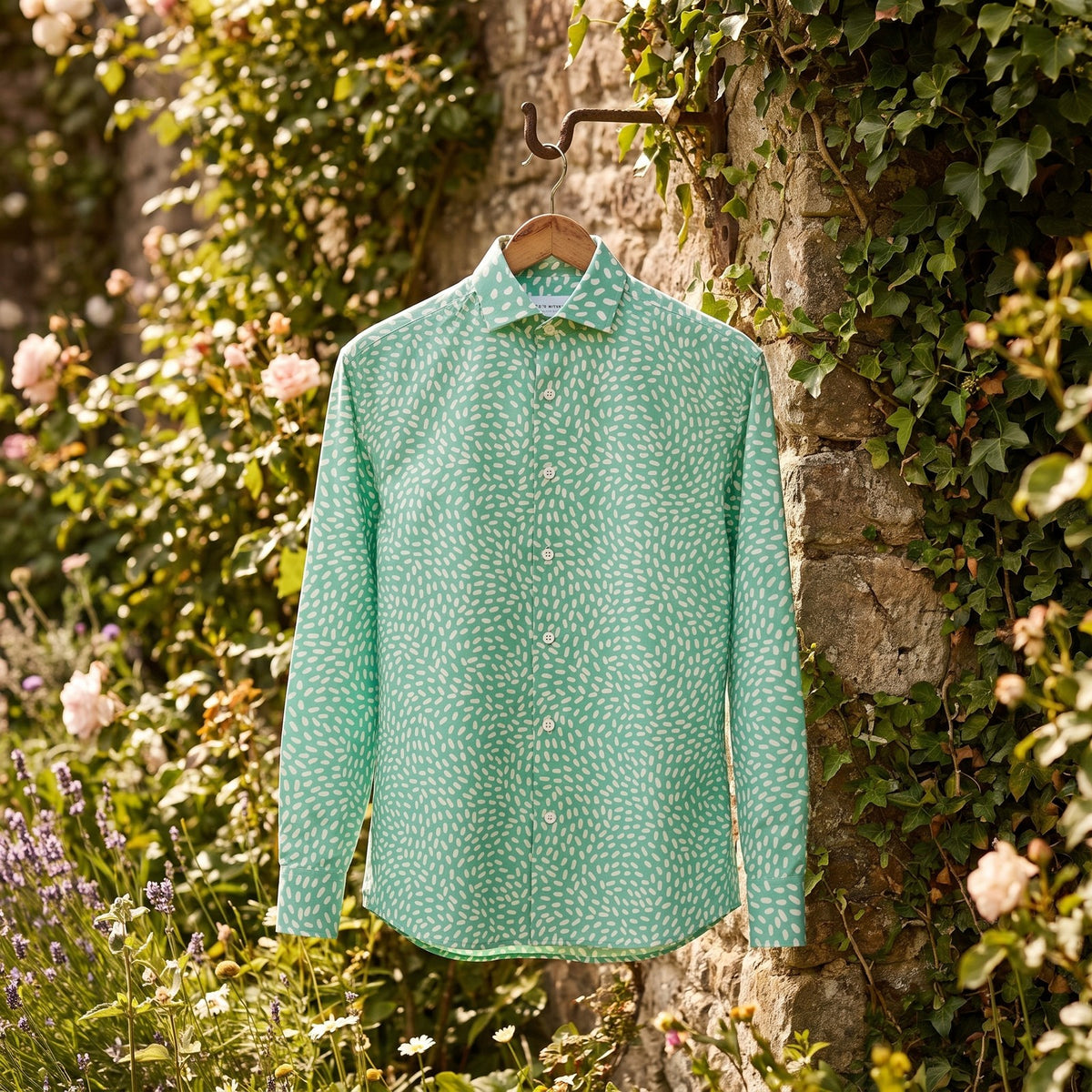 Chemise homme à manches longues vert menthe à petites touches blanches suspendue sur un cintre, à un crochet sur un vieux mur de pierre couvert de lierre dans un jardin fleuri - CHT'M Boutique