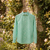 Chemise homme à manches longues vert menthe à petites touches blanches suspendue sur un cintre, à un crochet sur un vieux mur de pierre couvert de lierre dans un jardin fleuri - CHT'M Boutique