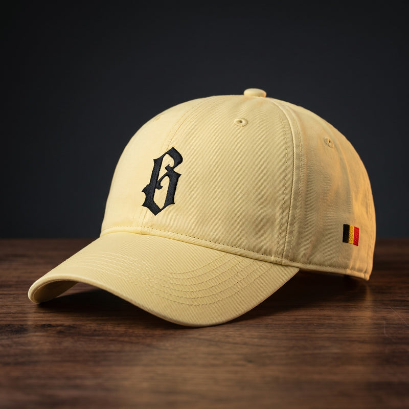 Casquette belge originale brodée B gothique jaune beurre, posée sur bois foncé, vue trois-quarts avec drapeau belge