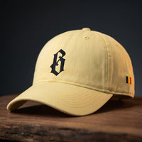 Dad cap belge délavée jaune beurre brodée B gothique, vue basse sur support bois foncé
