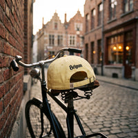 Casquette belge jaune beurre brodée Belgium au dos, posée sur la selle d'un vélo vintage dans une rue pavée de Bruxelles