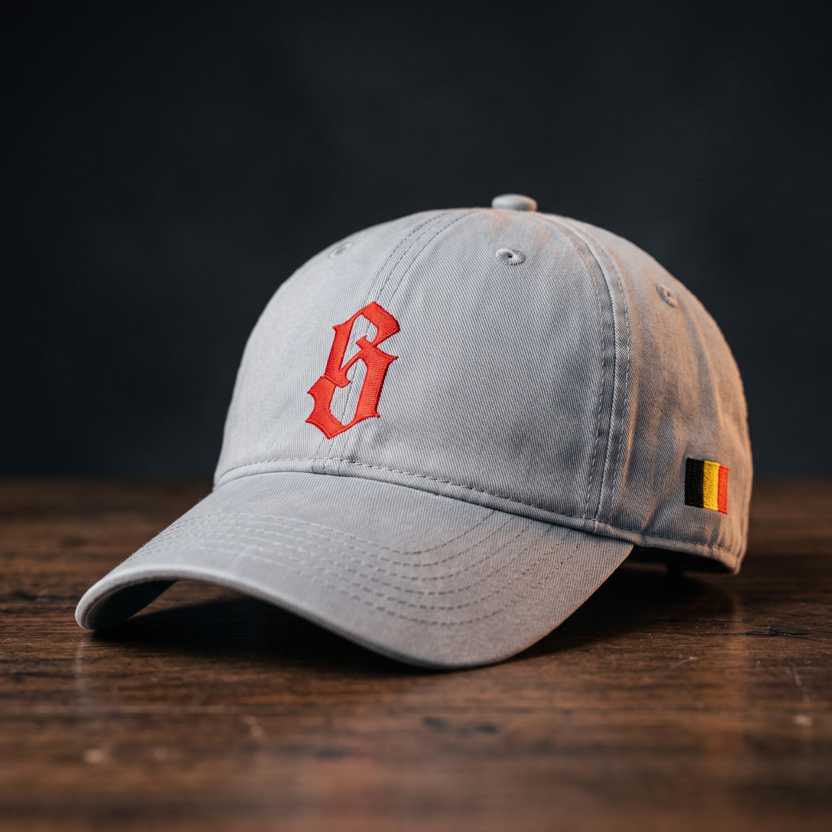 Casquette belge originale brodée B gothique rouge, coloris gris granit, posée sur bois foncé, vue trois-quarts