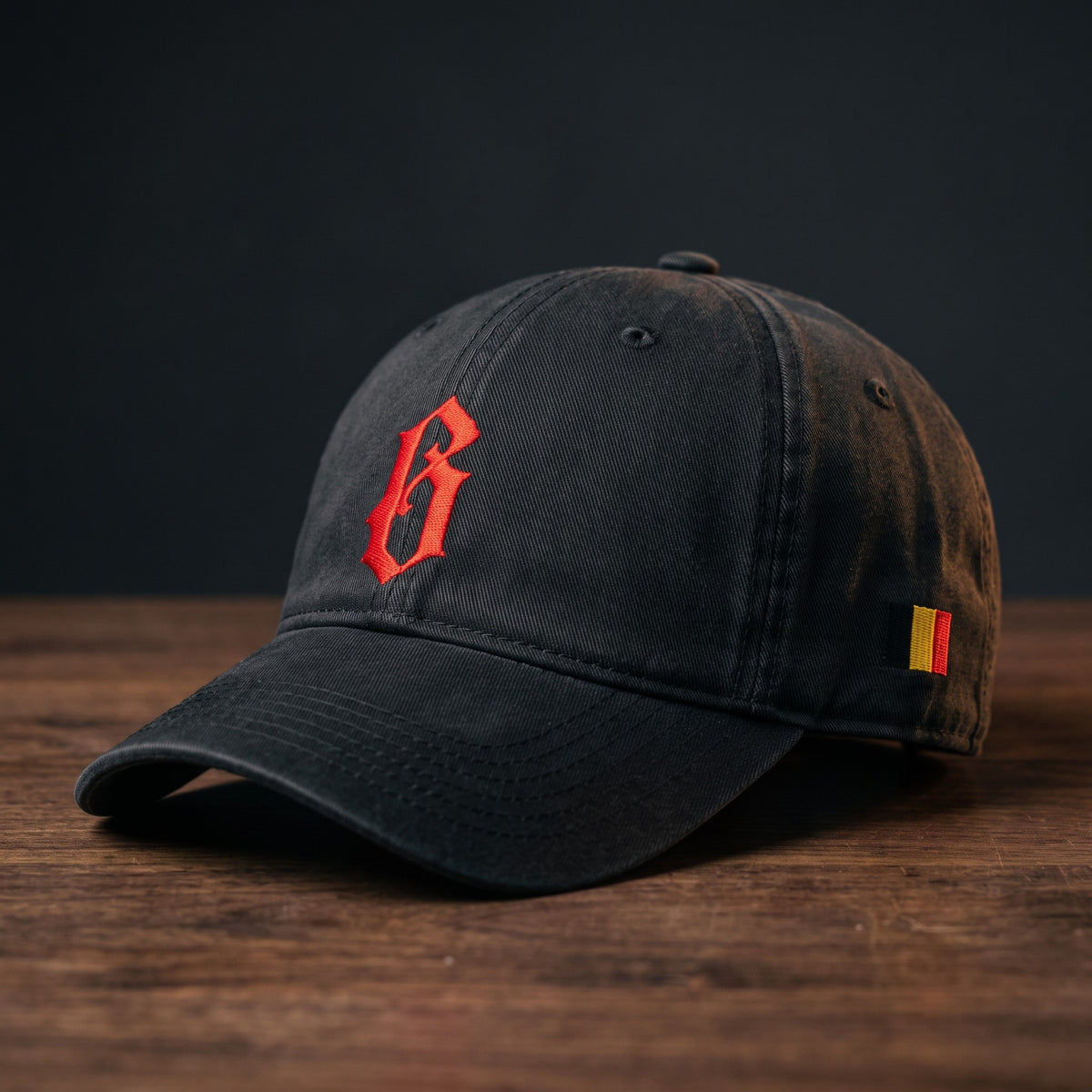 Casquette belge originale brodée B gothique rouge, coloris noir, posée sur bois foncé, vue trois-quarts avec drapeau belge