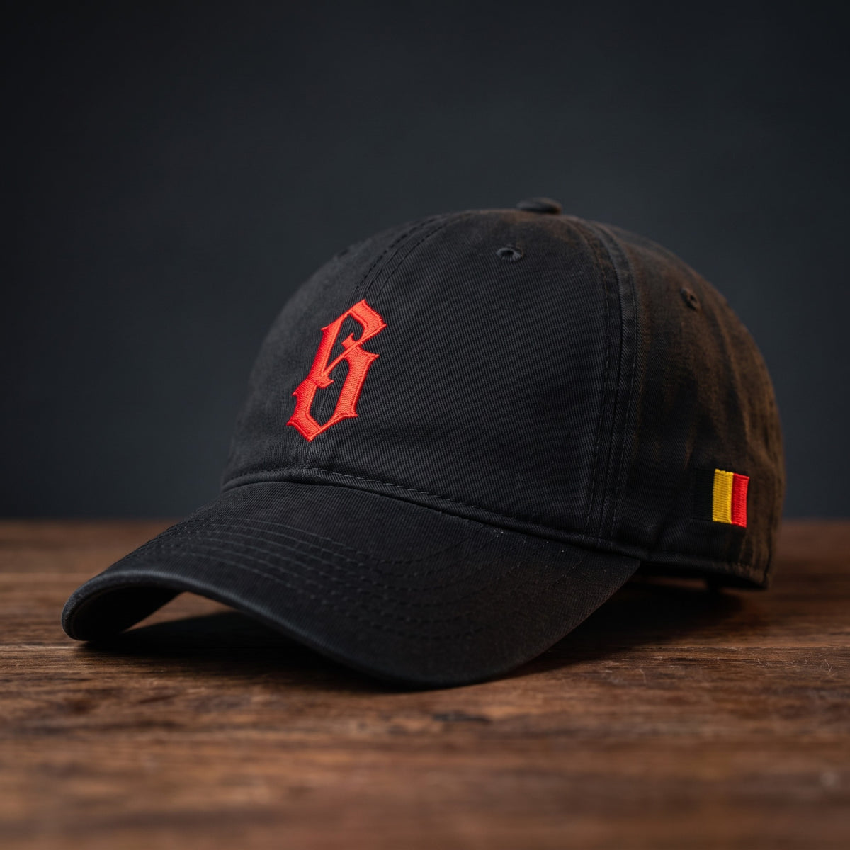Dad cap belge délavée noire brodée B gothique rouge, vue basse sur support bois foncé