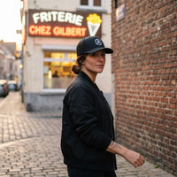 Casquette Goonies Never Say Die noire portée par une femme devant une friterie belge
