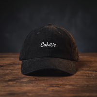 Casquette en velours côtelé noire brodée « Calvitie », posée sur une table en bois sombre, mise en scène cinématographique CHT'M Boutique.