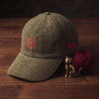 Casquette en velours côtelé olive CHT'M Boutique brodée tête de chat géométrique low-poly rouge et texte Cht'm gothique rouge, mise en scène lifestyle avec figurine de chat en laiton et pelote de laine rouge