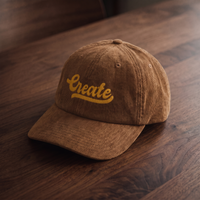 Casquette en velours côtelé brun avec broderie 'Create' dorée, vue trois-quarts sur bois de noyer foncé