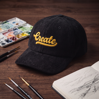 Casquette dad cap en velours côtelé noir CREATE 3D PUFF mise en scène dans un atelier d'artiste, entourée de palette d'aquarelle, pinceaux et carnet de croquis ouvert