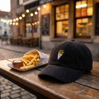 Casquette belge en velours côtelé noire brodée frites, posée sur une table de rue belge avec un cornet de frites