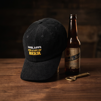 Casquette en velours côtelé noir avec broderie « Luck, Love and a lot of Beer », posée dans une mise en scène avec bouteille de bière vintage et décapsuleur en laiton, sur bois de noyer foncé