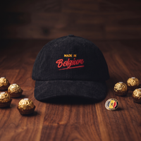 Casquette en velours côtelé noire Made in Belgium posée avec pralines belges et pin's émaillé belge sur bois foncé