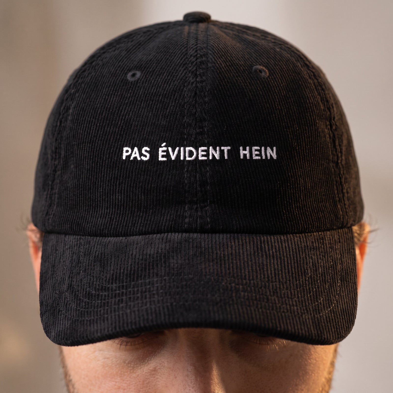 Gros plan de la broderie blanche « Pas évident hein » sur la casquette velours côtelé noire portée