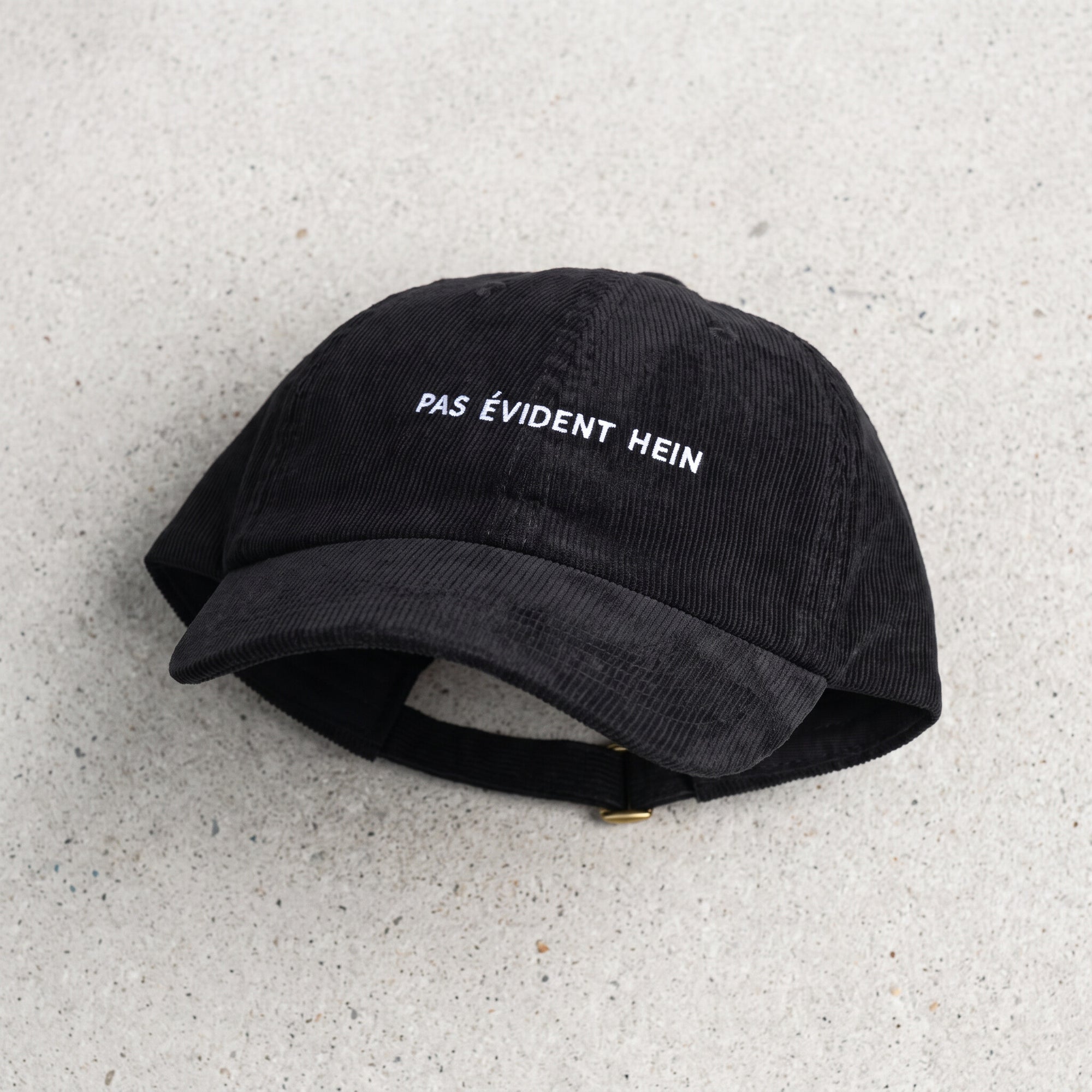 Casquette dad hat velours côtelé noire « Pas évident hein » posée à plat, design complet
