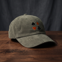 Casquette en velours côtelé olive avec quatre symboles de jeu de cartes brodés (pique, carreau, cœur, trèfle), posée sur bois foncé