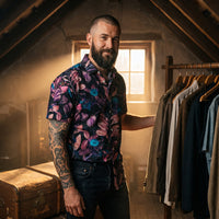 Chemise homme manches courtes noire à feuillage rose et fleurs bleues portée par un homme dans un grenier vintage près d'un portant et d'une malle en cuir - CHT'M Boutique