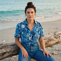 Chemise homme manches courtes bleue à fleurs blanches et feuillage marine portée par une femme assise sur un tronc d'arbre échoué sur une plage - CHT'M Boutique