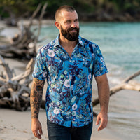 Chemise homme manches courtes bleue à fleurs blanches et feuillage marine portée par un homme marchant sur une plage calme au bord de l'eau turquoise - CHT'M Boutique