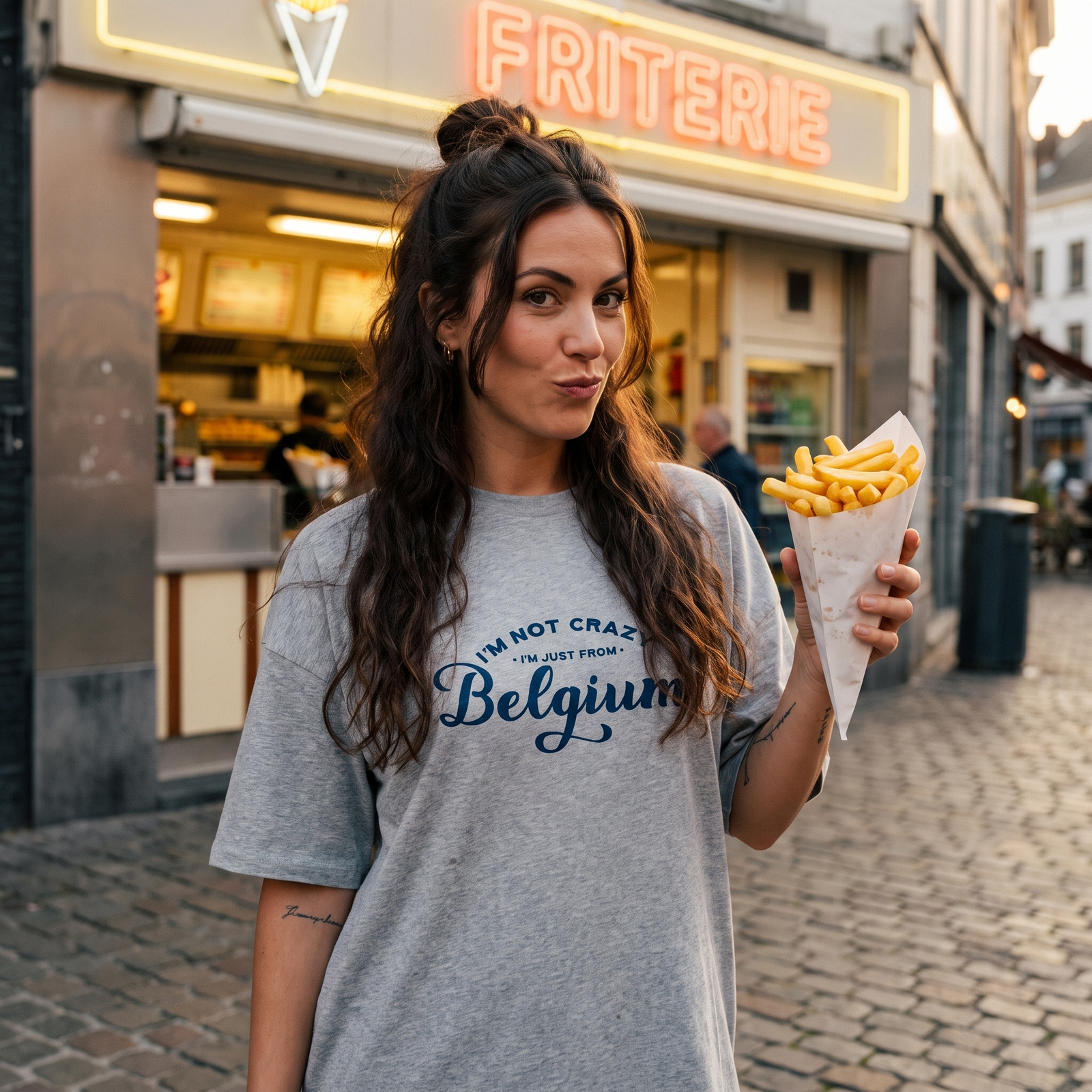 Robe t-shirt oversize grise avec inscription humoristique "I'm not crazy, I'm just from Belgium", portée devant une friterie belge illuminée