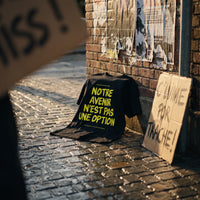 T-shirt Gen Z noir « Notre avenir n'est pas une option » posé sur les pavés après une manif, pancarte en carton contre le mur – t-shirt manif original CHT'M