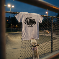 T-shirt Gen Z gris avoine « Stay Strong la Gène Z » accroché au grillage d'un skatepark au crépuscule – tshirt genz pas cher CHT'M