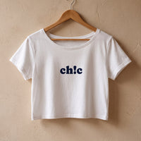 Crop top femme blanc imprime ch!c suspendu sur un cintre en bois clair devant un mur de platre beige