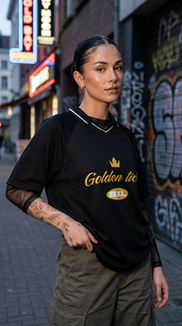 Femme portant le maillot Golden Tich noir en rue – mur graffiti, style streetwear, design doré BXL