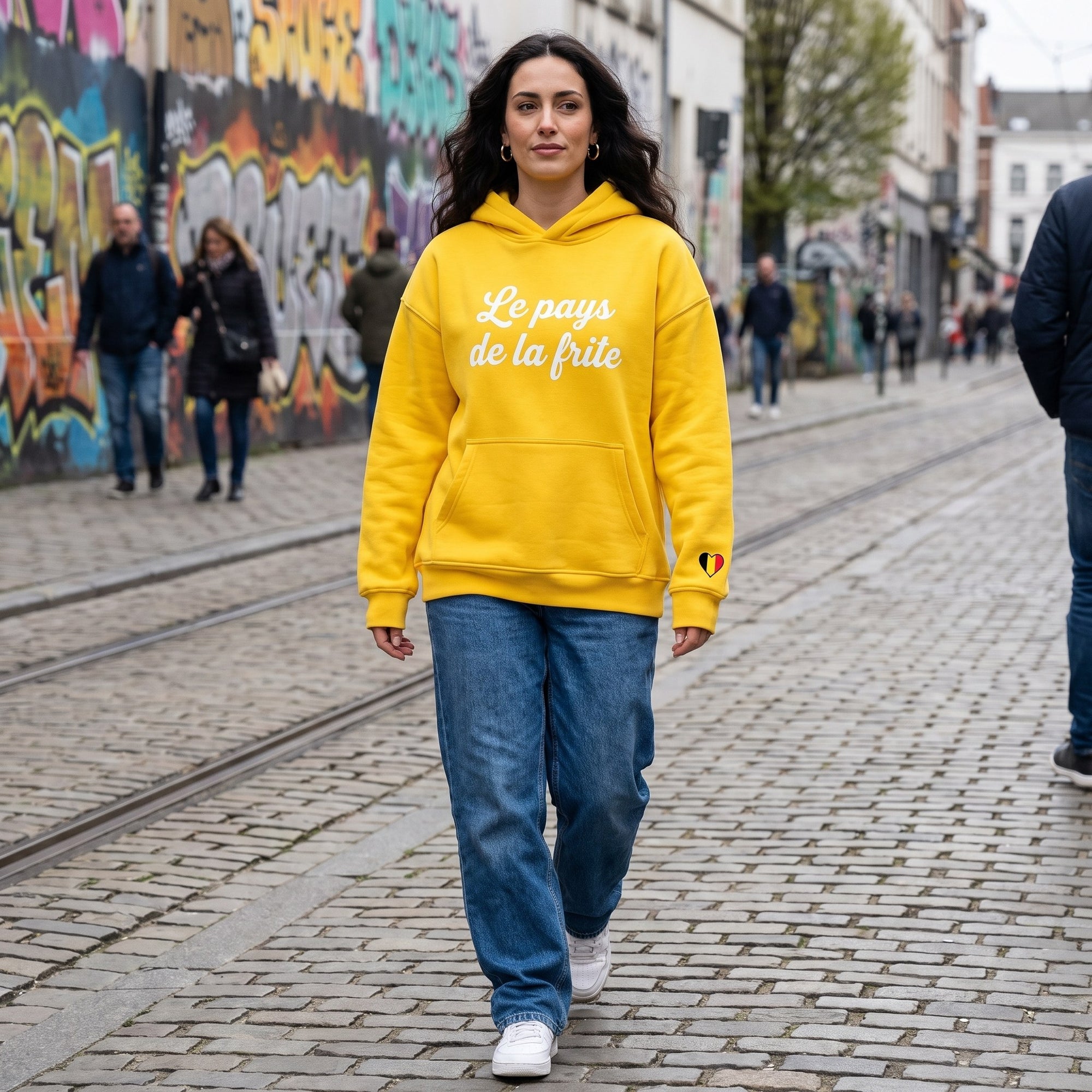 Femme en hoodie belge original Le Pays de la Frite jaune dans une rue belge, look streetwear oversize