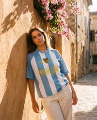 Femme portant le maillot streetwear Wesh Pélo Bleu Ciel & Blanc dans une ruelle fleurie de bougainvilliers