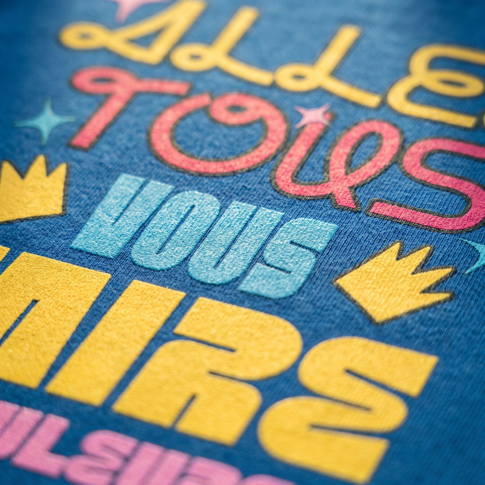 Gros plan sur le lettrage jaune, rose et bleu Allez tous vous faire en couleurs imprimé sur le coton bleu du t-shirt