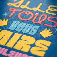 Gros plan sur le lettrage jaune, rose et bleu Allez tous vous faire en couleurs imprimé sur le coton bleu du t-shirt