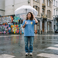 Femme sous un parapluie transparent en rue portant le t-shirt belge bleu Né sous une drache d'amour