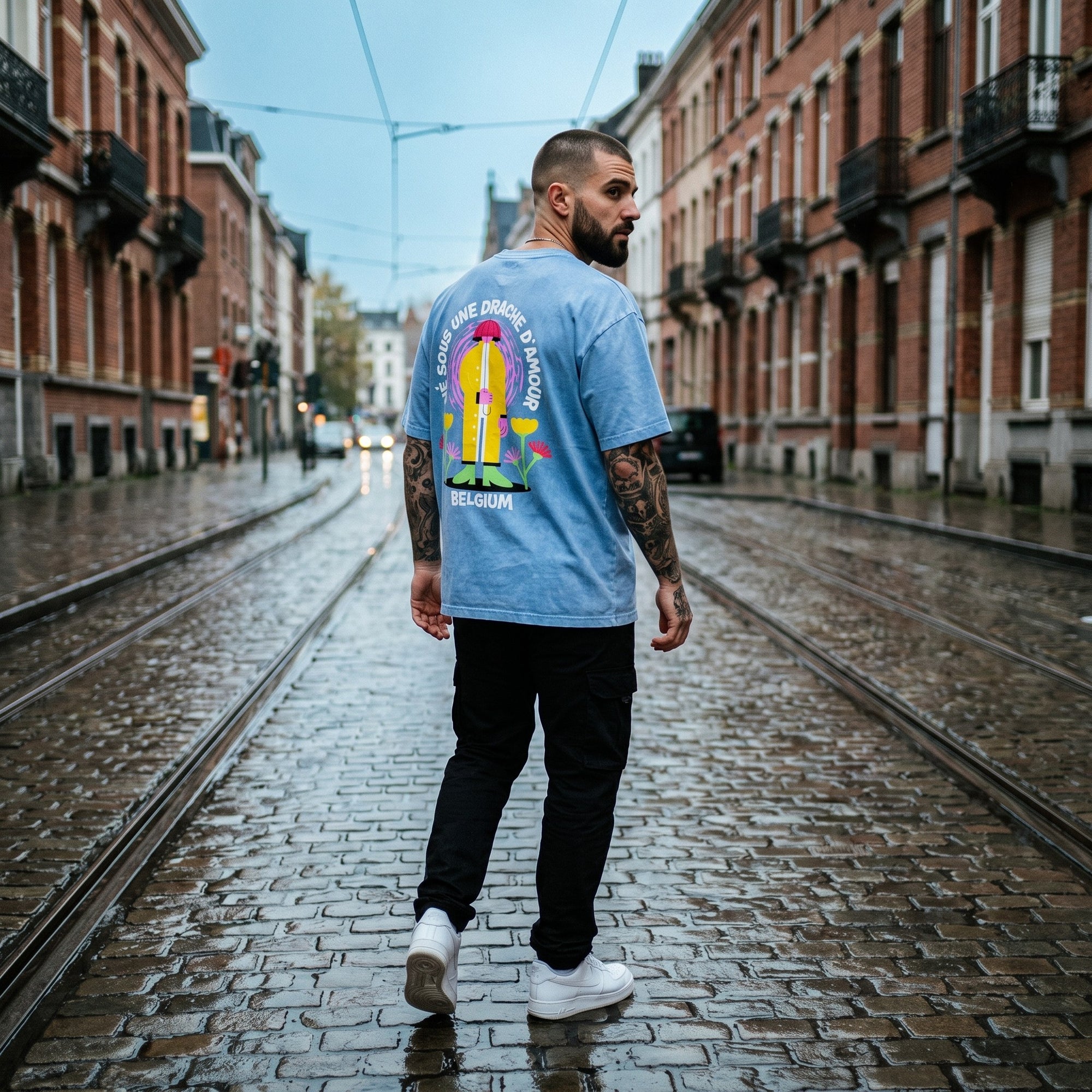 Streetwear belge : homme tatoué portant le t-shirt belge original Né sous une drache d'amour dans une rue pavée de Bruxelles sous la pluie