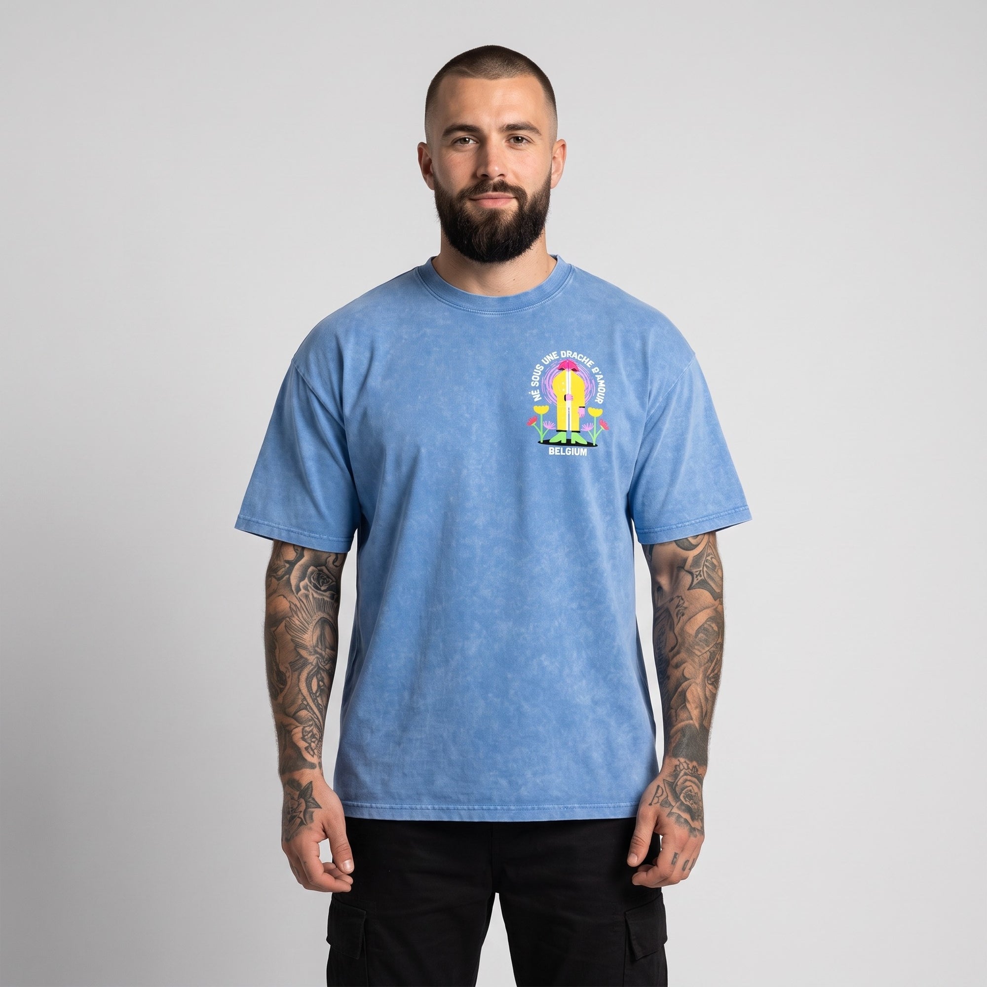 Homme buzz cut, barbe et tatouages manche portant le t-shirt belge original Né sous une drache d'amour, coupe oversize bleu délavé, photo studio de face