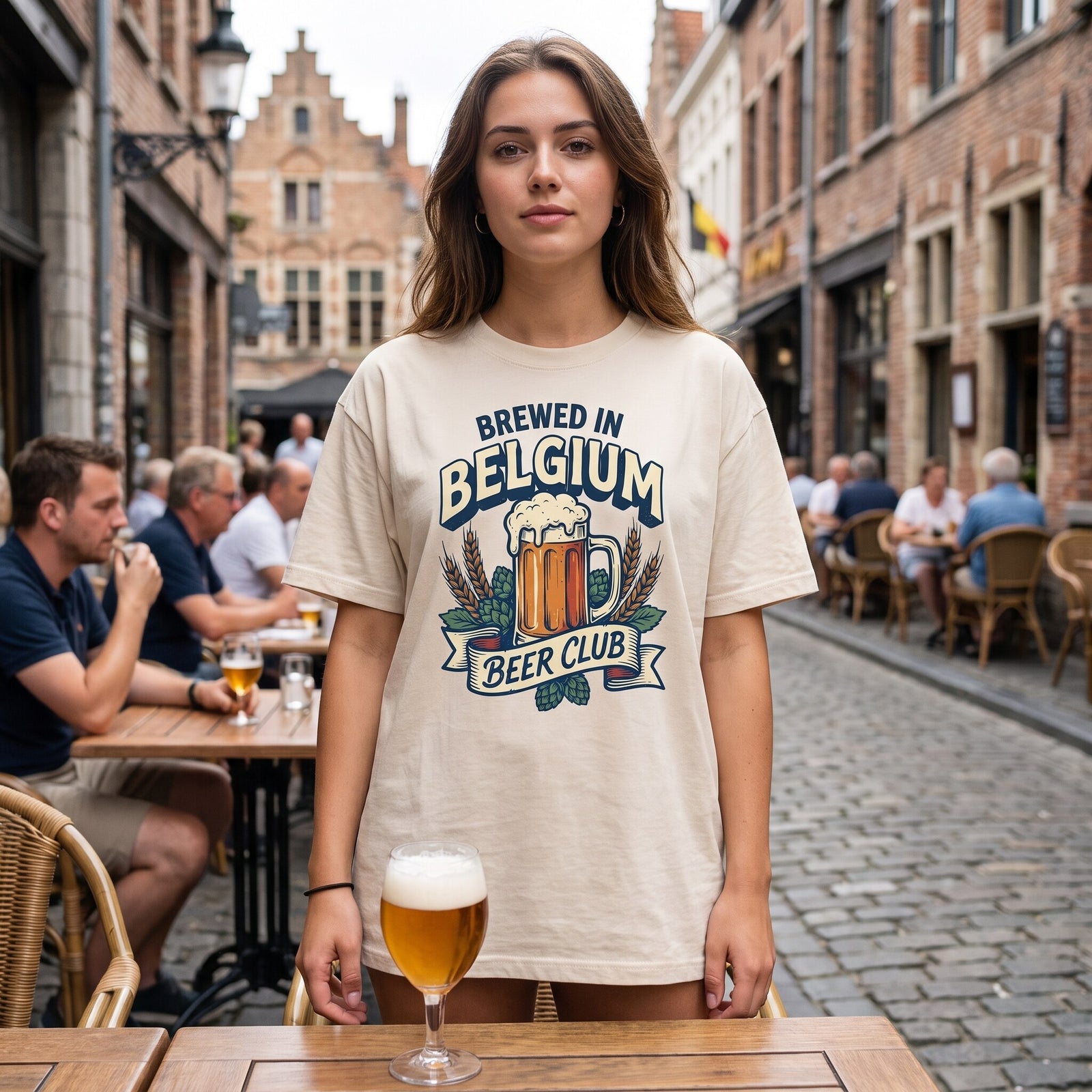 Femme en terrasse d'une ruelle pavée belge, t-shirt écru Brewed in Belgium Beer Club et verre de bière posé sur la table