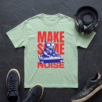 T-shirt vert clair imprimé chat DJ rouge et bleu, posé à plat sur une ardoise noire avec un casque audio et des sneakers