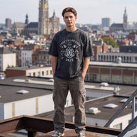 T-shirt oversized noir lavé à tête de chat blanche, plein pied sur une poutre avec les toits d'Anvers derrière