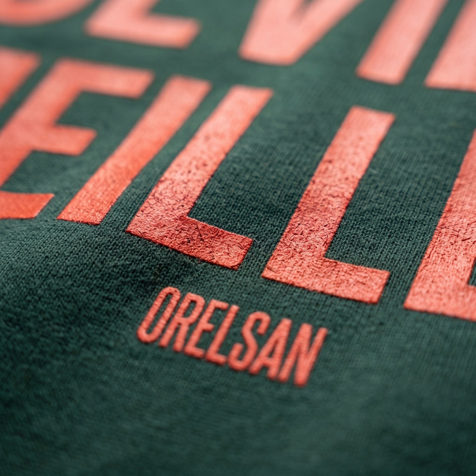 Gros plan sur le lettrage orange Deviens Meilleur et la signature Orelsan imprimés sur le coton vert forêt