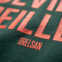 Gros plan sur le lettrage orange Deviens Meilleur et la signature Orelsan imprimés sur le coton vert forêt