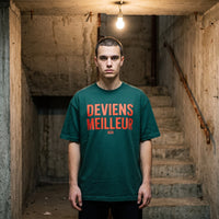 T-shirt oversized vert forêt Deviens Meilleur, porté dans une cage d'escalier en béton brut éclairée par une ampoule nue