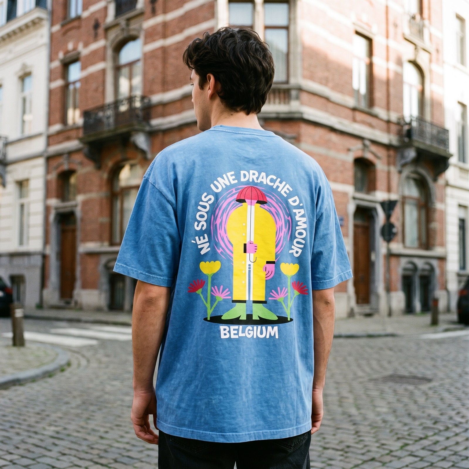 Homme de dos en t-shirt bleu snow wash Né sous une drache d'amour, à un coin de rue bruxellois aux façades de brique
