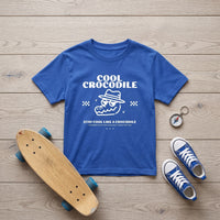 T-shirt enfant bleu vif au crocodile à chapeau, posé à plat sur un parquet clair avec skateboard et baskets bleues