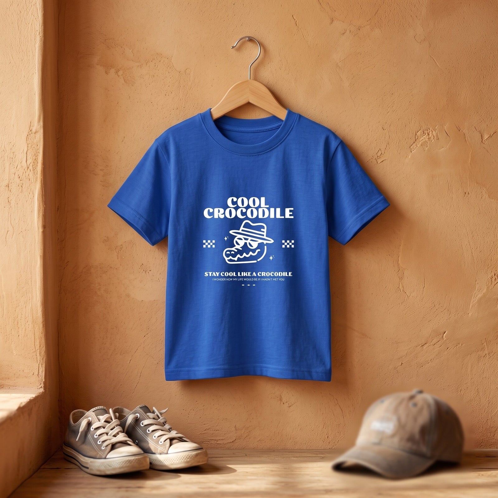 T-shirt enfant bleu vif Cool Crocodile sur cintre en bois devant un mur ocre, baskets et casquette posées au sol