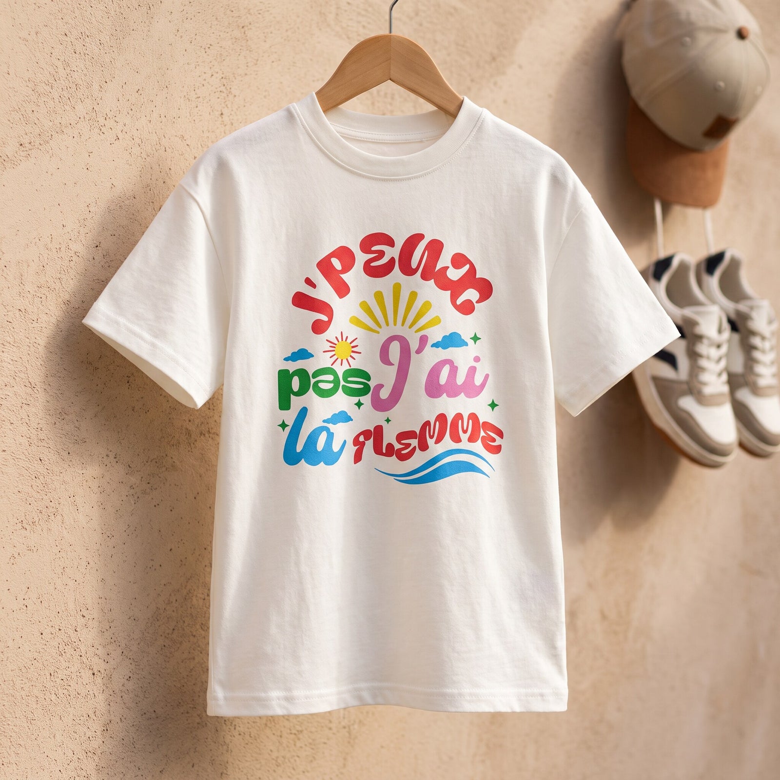 T-shirt enfant blanc a l'imprime multicolore sur cintre, casquette beige et sneakers accrochees au mur de sable